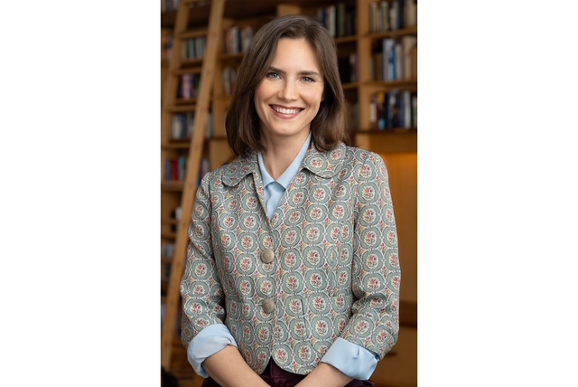 A woman with brown hair smiles warmly, wearing a patterned jacket over a light blue shirt. She stands in front of a bookshelf filled with various books.