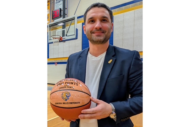 A man in a basketball court holds a commemorative ball celebrating 1,000 career points. He's smiling, wearing a blazer, giving a proud, happy vibe.