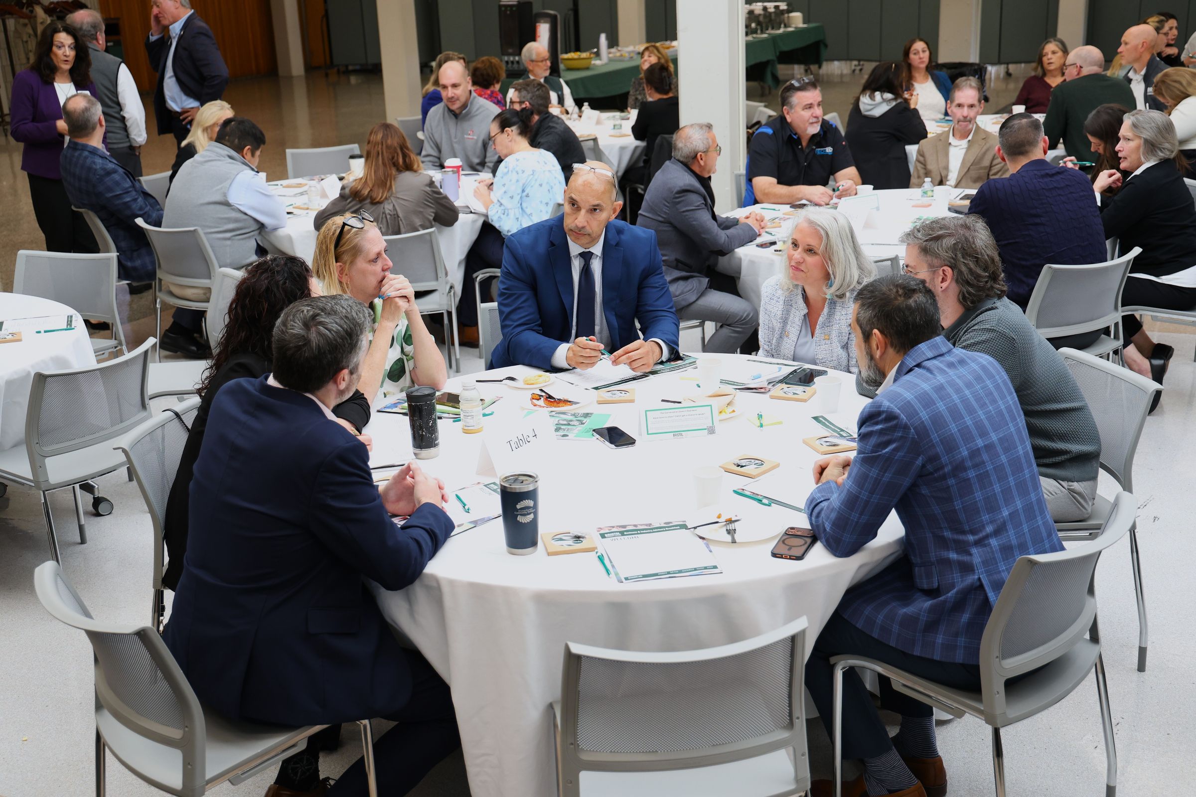 Professionals gather around round tables in a bright conference room, discussing papers and sharing ideas in a collaborative, focused workshop.