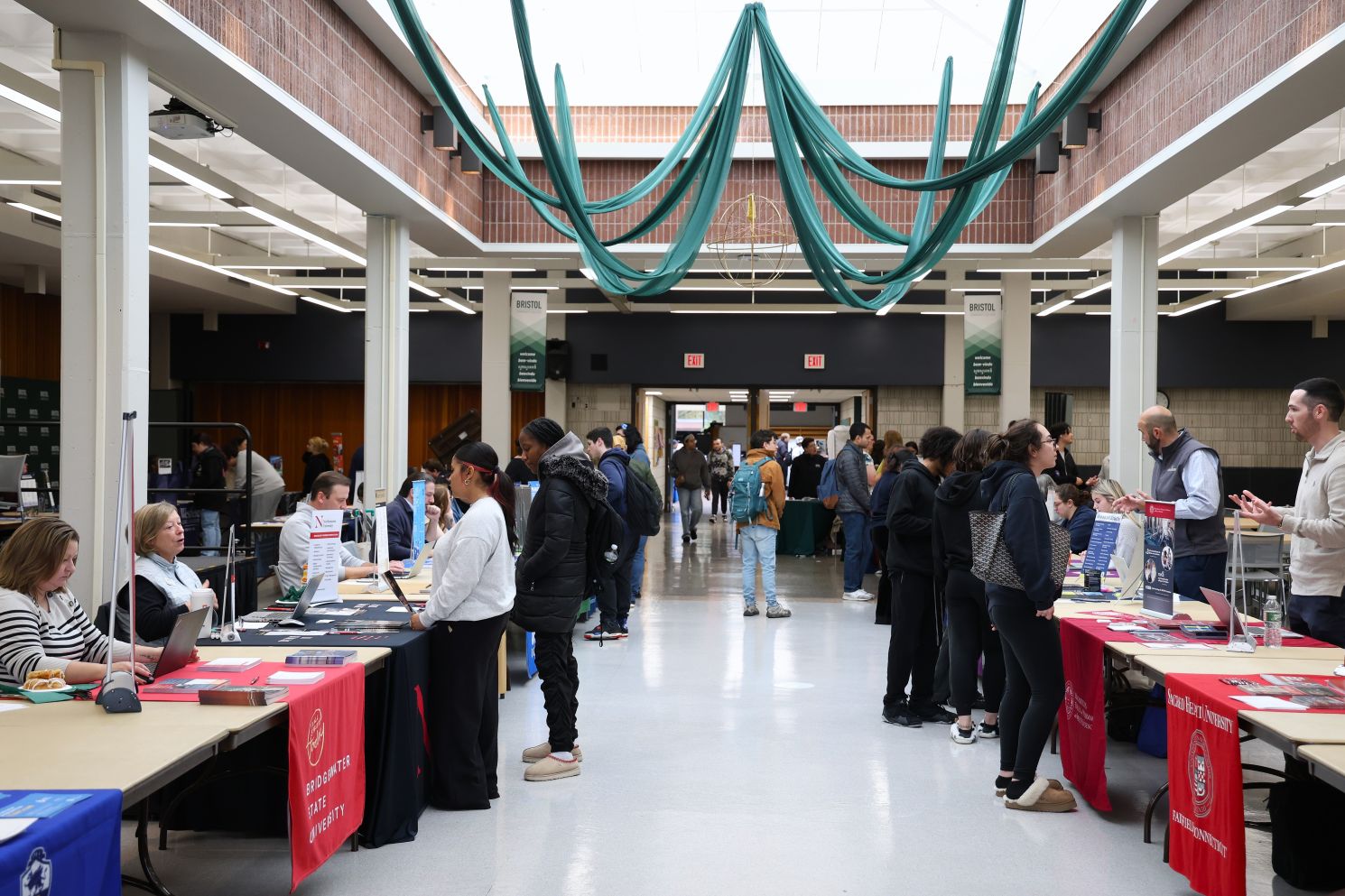 Visitors browse information tables at a busy campus fair in a bright atrium, beneath looping teal fabric banners.