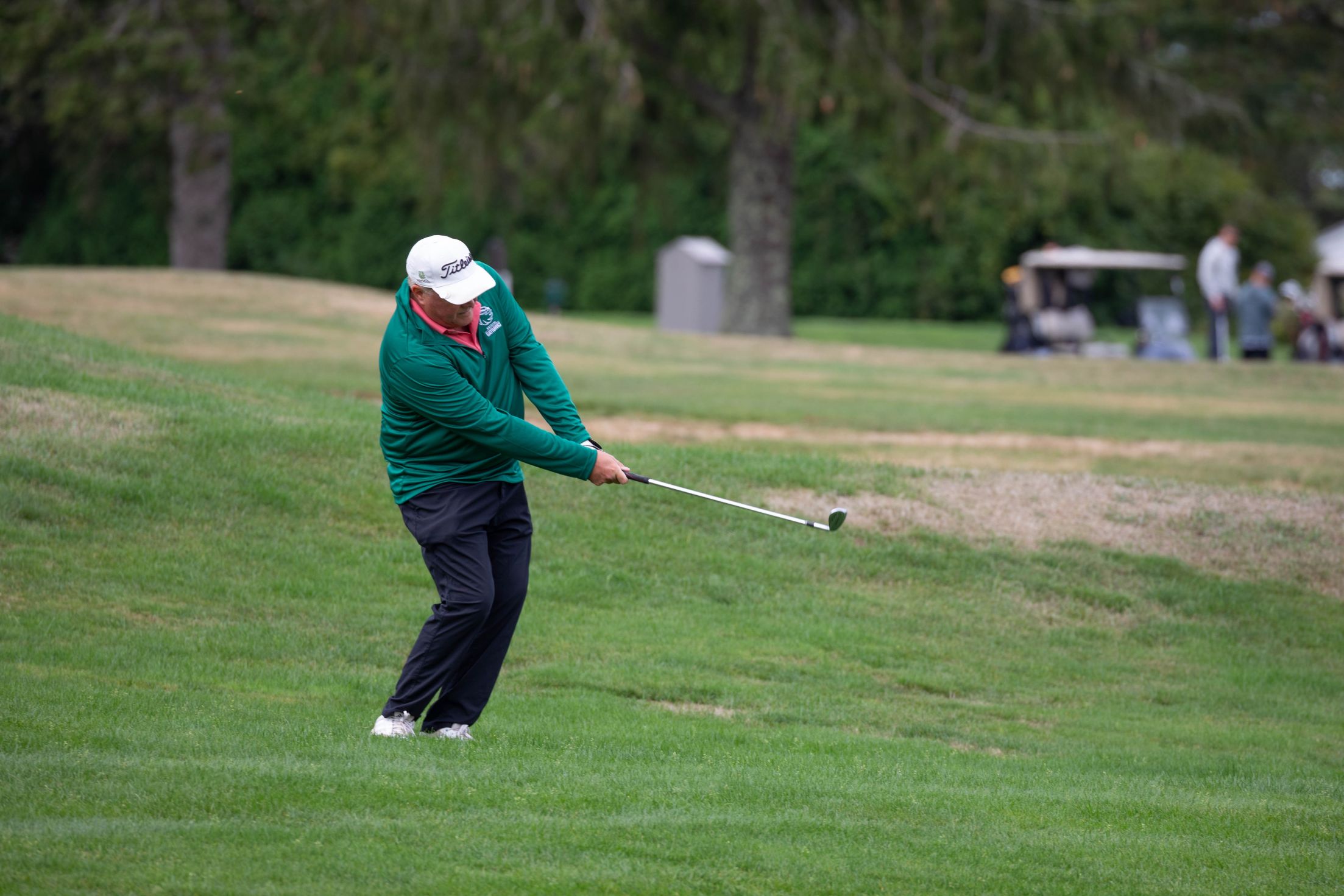 A golfer in a green jacket swings a club on a grassy course, with trees and golf carts visible in the background.