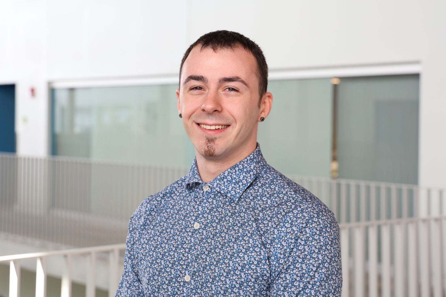 A person smiles warmly while standing indoors, wearing a blue floral shirt. The modern, light-filled space includes glass and metal accents, conveying a professional and friendly atmosphere.