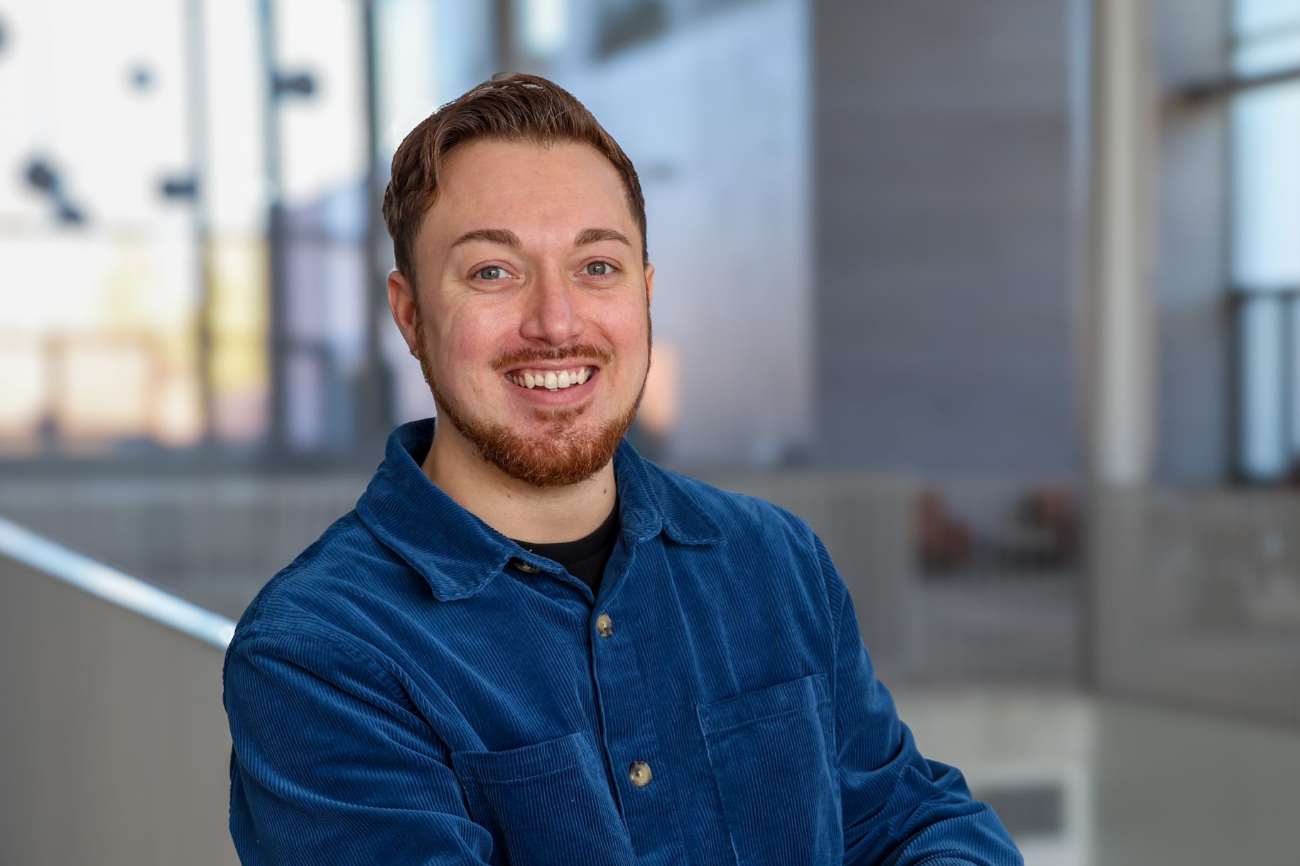 A smiling person with short brown hair and a beard, wearing a blue corduroy shirt, stands indoors with a blurred modern background.