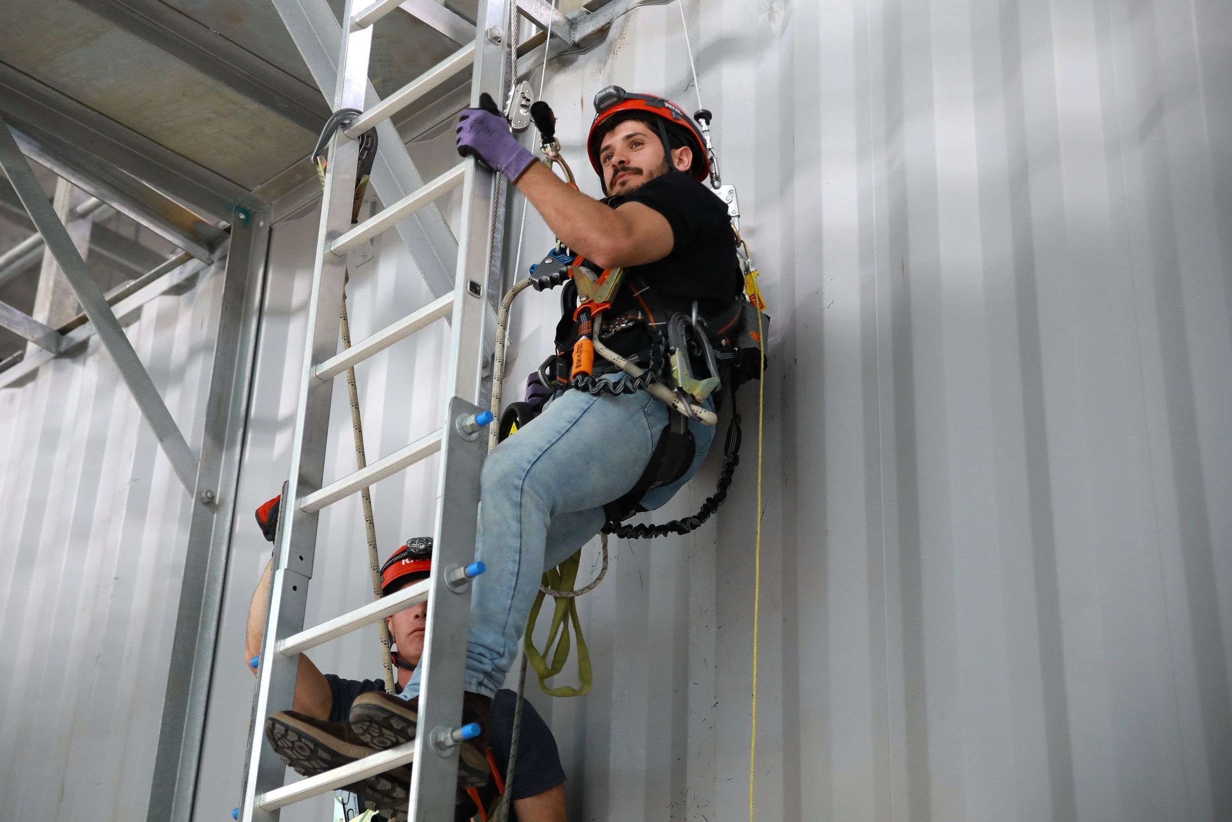 Rope-access technician in a red helmet and safety harness climbs a ladder inside a metal structure, focused during industrial work.