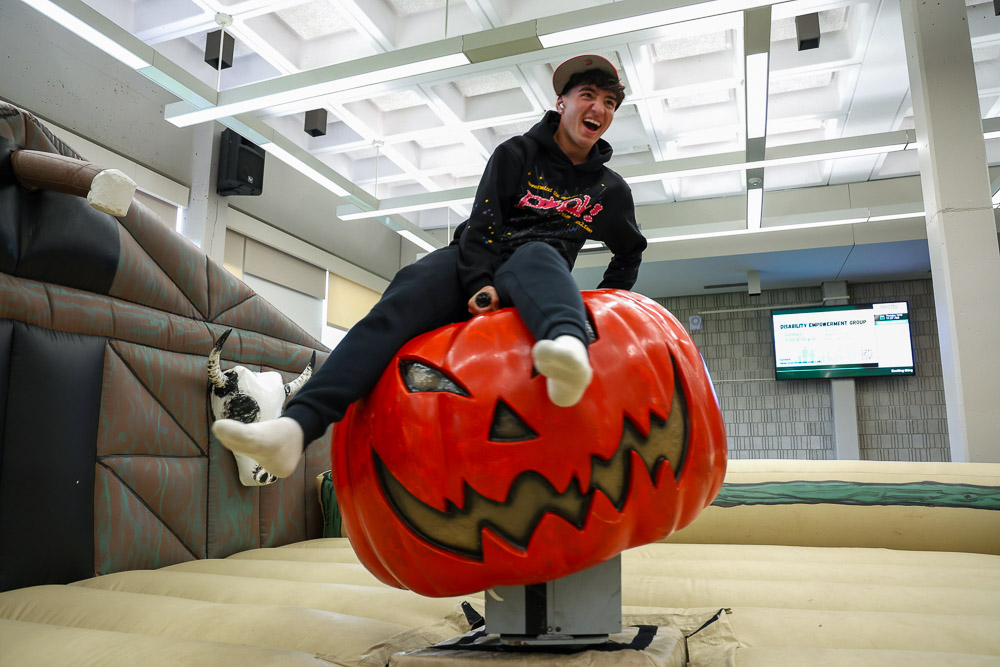 Student riding a mechanical bull style machine. The machine is styled as a jack-o-lantern.